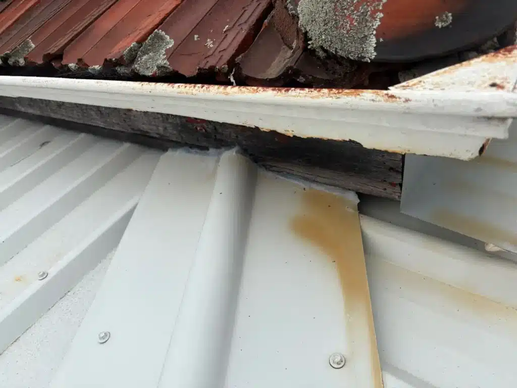 Rusted gutter and water staining at the junction of tiled and metal roofing, showing a potential source of roof leaks.